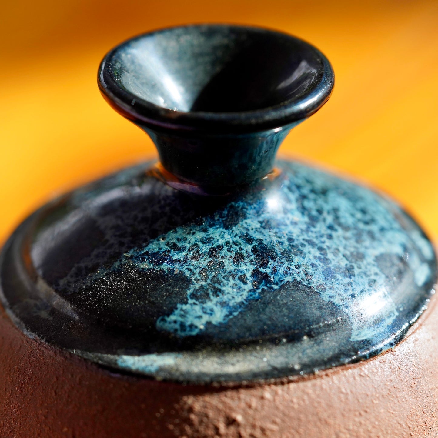 Macro photo of of black, white, and blue nebula glaze on top of a handmade brown clay ceramic bud vase, displayed on a light wood floor background