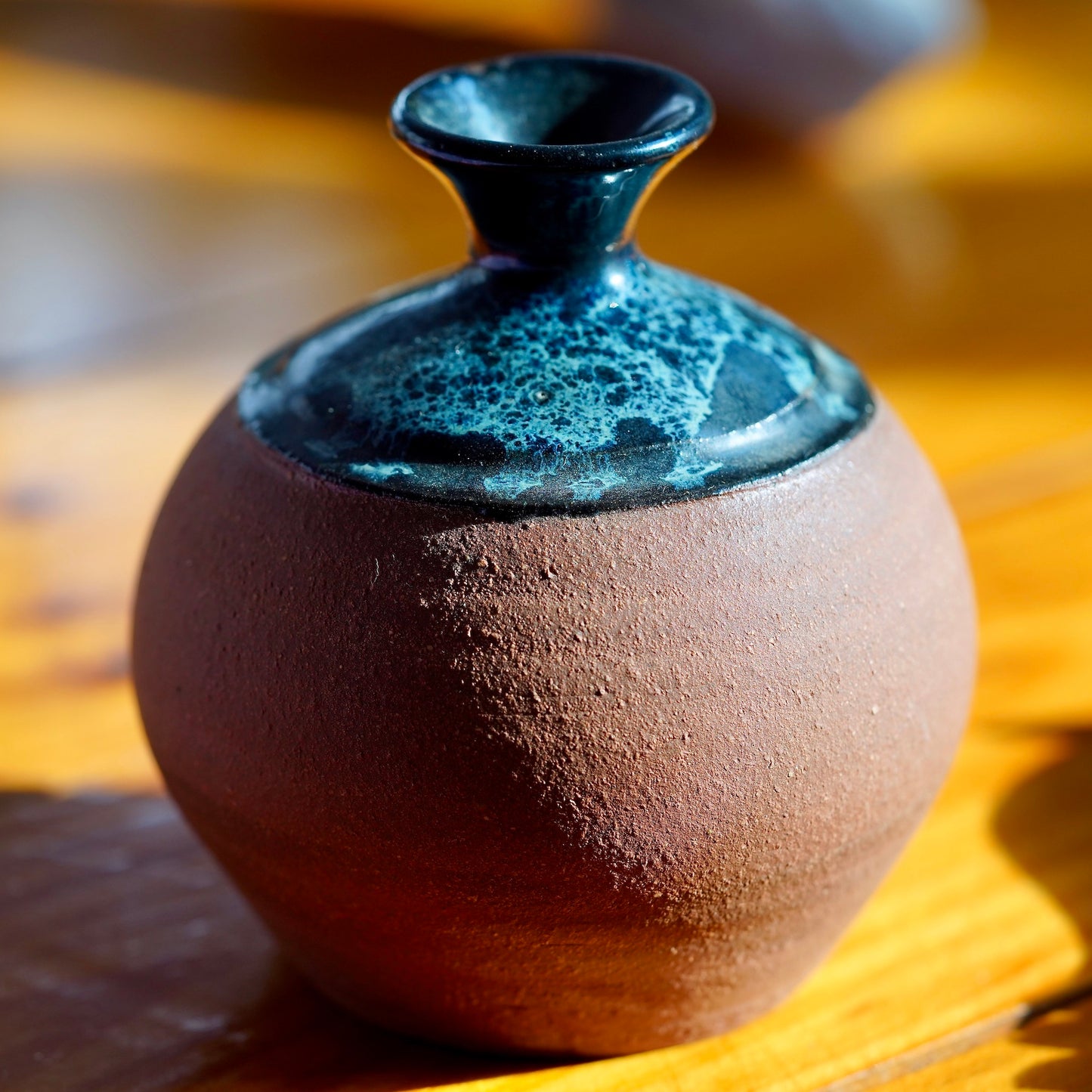 Handmade brown clay ceramic bud vase with black, blue and white nebula glaze on top, displayed on a light wood floor